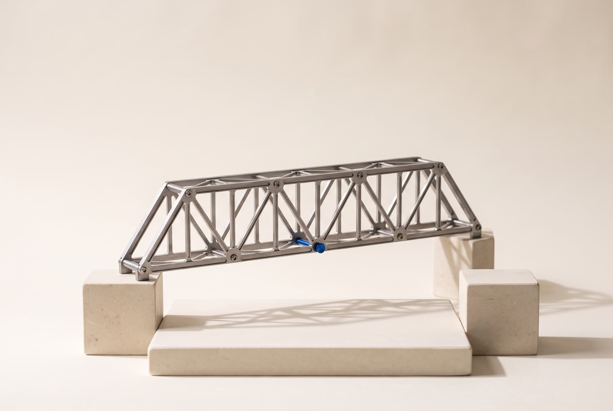 A precision model of a steel truss bridge spanning two stone blocks, one cobalt pin at its centre